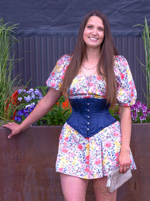 Cute corset model in a floral dress wearing the cs 201 satin hourglass curve corset in navy in front of a flower box