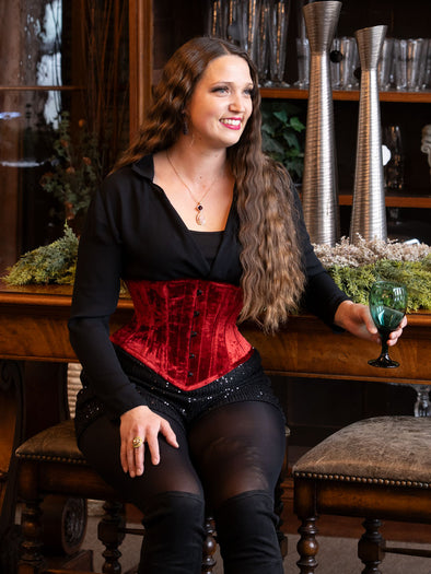 woman with long hair sitting in a lounge wearing a red velvet coset for waist training and fashion with sequin shorts and over the knee boots