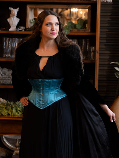 Woman wearing a black dress with a teal blue waspie corset belt in a room with decorative items.