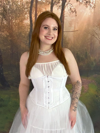 Model standing in fantasy forest wearing a white fairycore dress with a white waspie corset and pearls cs-220 waist training corset for weddings and boudoir shoots 