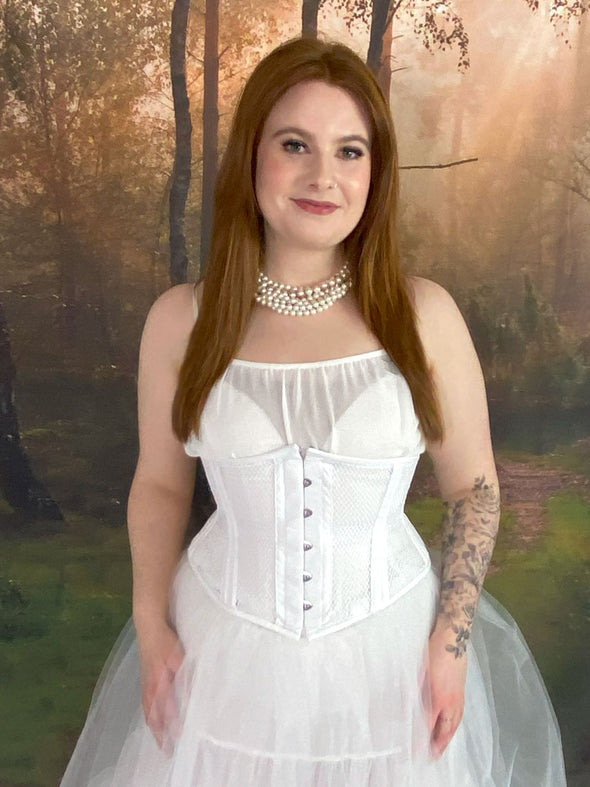 Model standing in fantasy forest wearing a white fairycore dress with a white waspie corset and pearls cs-220 waist training corset for weddings and boudoir shoots