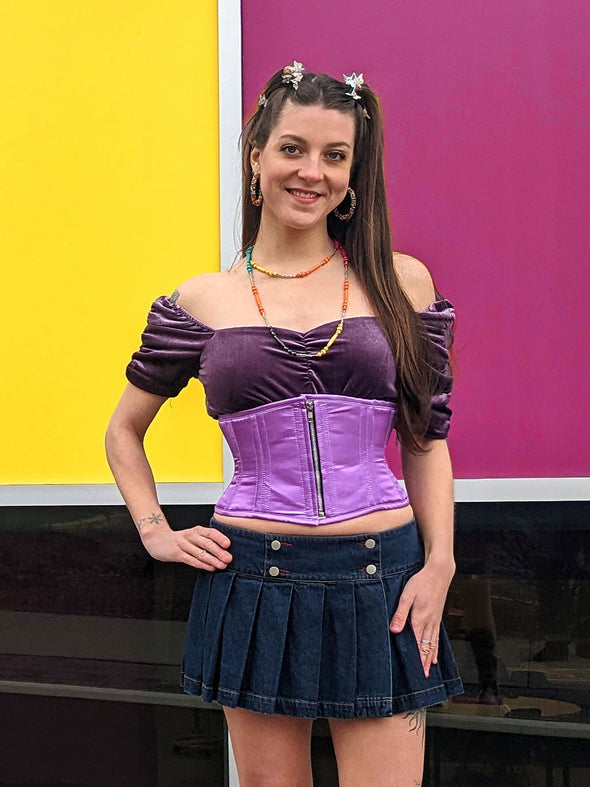 Sassy cute model wearing a short lilac corset belt with front zipper closure. Standing with one hand on her hip and wearing a short denim skirt and platform shoes