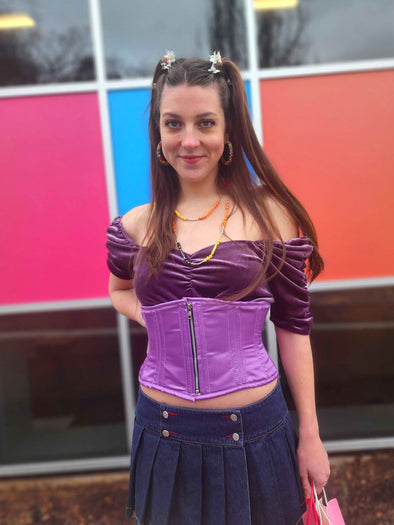 Sassy cute model wearing a short lilac corset belt with front zipper closure. Standing with one hand on her hip and wearing a short denim skirt and platform shoes