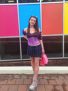 Sassy cute model wearing a short lilac corset belt with front zipper closure. Standing with one hand on her hip and wearing a velvet top, short denim skirt and platform shoes
