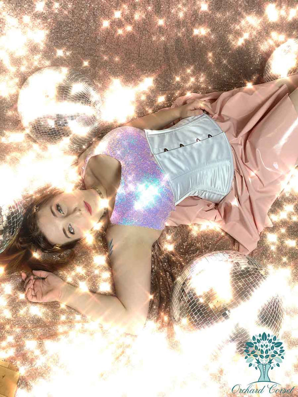 woman modeling a white satin romantic curve cs411 standard corset with pink latex skirt and irridescent strap blouse.  Laying on sequins with disco balls