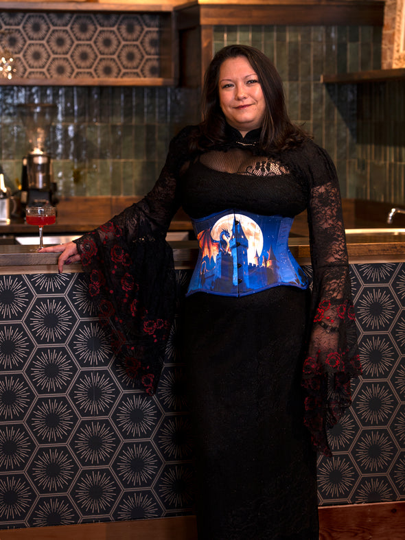 Woman wearing a black lace top and blue corset with a castle design in a bar setting.