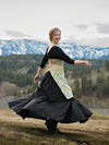 Woman in a black dress with a floral apron and beige corset standing in a scenic outdoor setting with mountains and trees.
