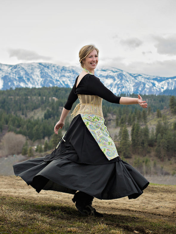 Woman in a black dress with a floral apron and beige corset standing in a scenic outdoor setting with mountains and trees.