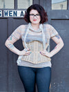 Woman wearing a beige mesh corset over a mesh top with a gray scarf over a gray background 