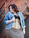 Woman wearing a denim jacket, black top, with a beige corset and striped skirt against a brick wall.
