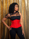 Model with dark curly hair wearing a festive Holiday red corset with a sheer top and black pants looking away from the camera