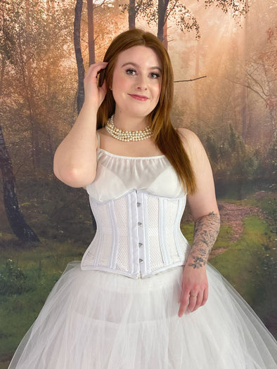 Model standing in a fantasy forest in a white fairycore dress with a white corset cs-426 Hourglass corset