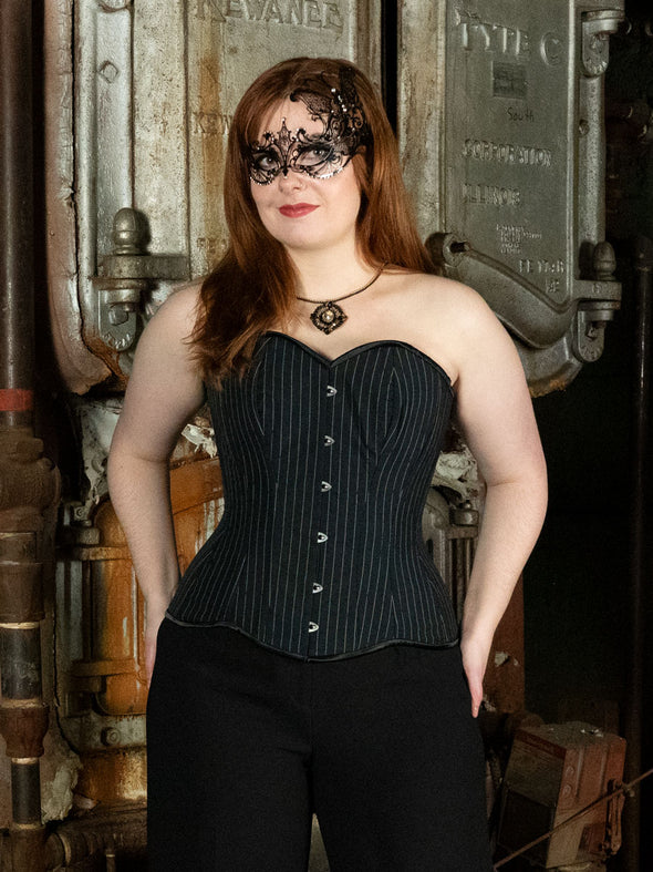 glamourous model wearing a pinstripe hourglass curve overbust corset top with black pants and a sparkly pendant and mask