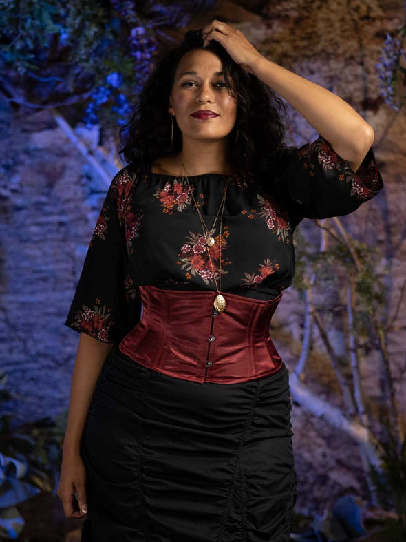 Model wearing the cs 301 waspie corset in wine colored silky satin.  Corset is worn over a dark floral blouse and black ruched cotton skirt.