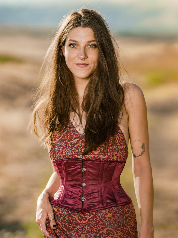 fresh faced model with long brown hair wearing a short wine colored satin corset over a floral dress