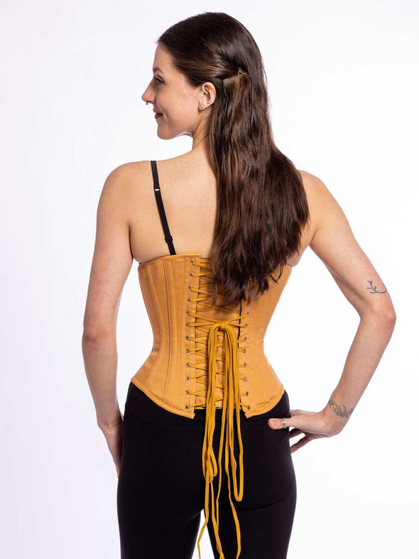 corset model wearing black tights and bra with a beige cotton everyday corset for fashion and waist training showing the back lace up view