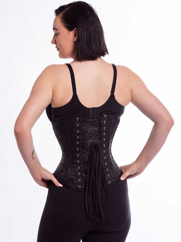 female model wearing the cs345 black brocade corset back lace up view with hands on hips
