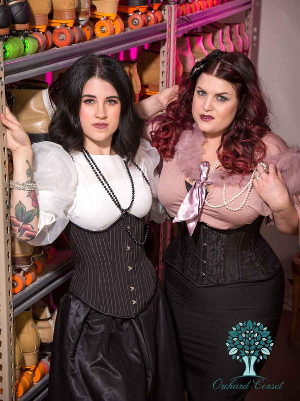 model wearing our underbust 345 pinstripe corset standing next to a model wearing our 426 black brocade corset