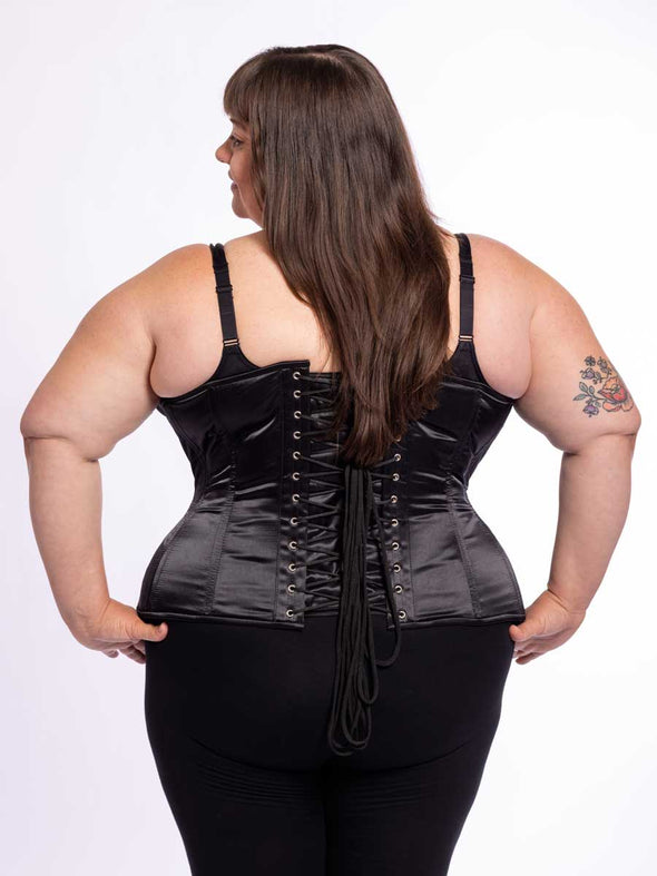 curvy plus sized model wearing black legging and bra with a black satin corset standing with her hands on her hips back lace up corset view