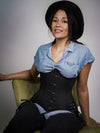 model sitting on a green velvet chair wearing jeans and a blue denim shirt with a black corset with hip ties and a black hat 