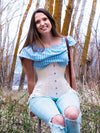 smiling model sitting on a ladder in a wooded ares wearing a striped ruffled blouse ripped jeans and boot and a long hourglass curve corset with hip ties to complete the outfit