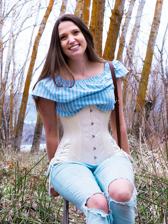 smiling model sitting on a ladder in a wooded ares wearing a striped ruffled blouse ripped jeans and boot and a long hourglass curve corset with hip ties to complete the outfit