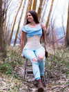 smiling model sitting on a ladder in a wooded ares wearing a striped ruffled blouse ripped jeans and boot and a long hourglass curve corset with hip ties to complete the outfit