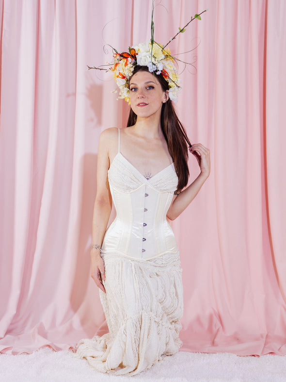 Model dressed in ivory wearing a slowing sundress, a flower crown and an ivory hourglass corset in front of a pink fabric backdrop