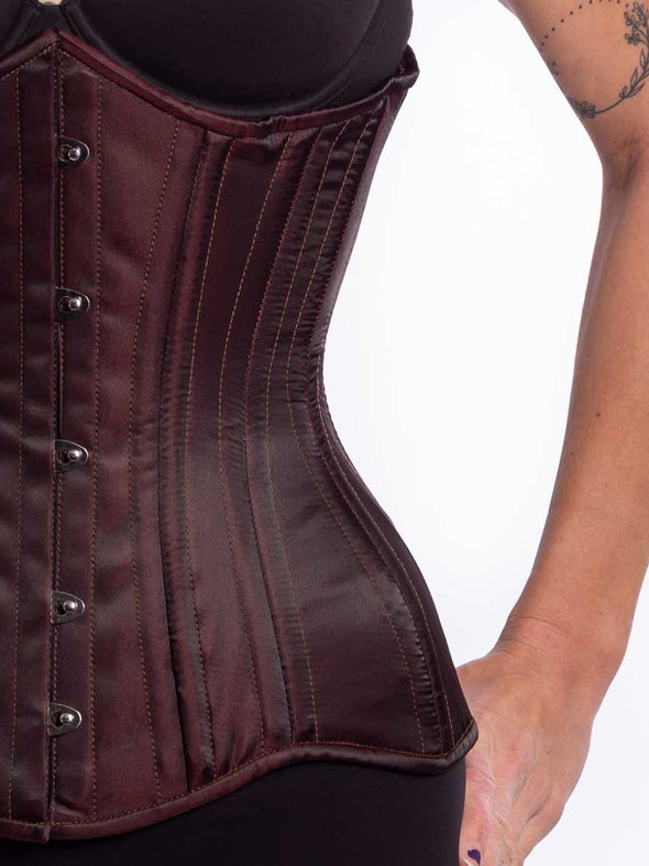 Front detail view of a Female model wearing a curvy longline matte maroon satin corset over black leggings and a black bra