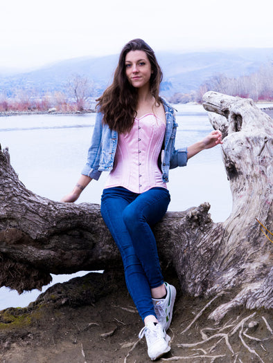 Model sitting on driftwood by a river wearing jeans and a faded jean jacket over a light pink corset top