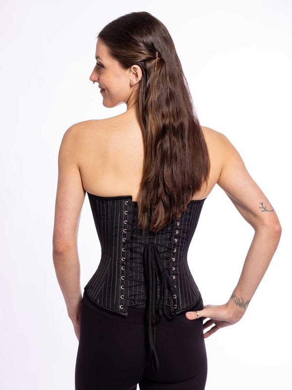 back lace up corset view of a cute corset model wearing an overbust strapless corset top in a black pinstripe fabric with black leggings
